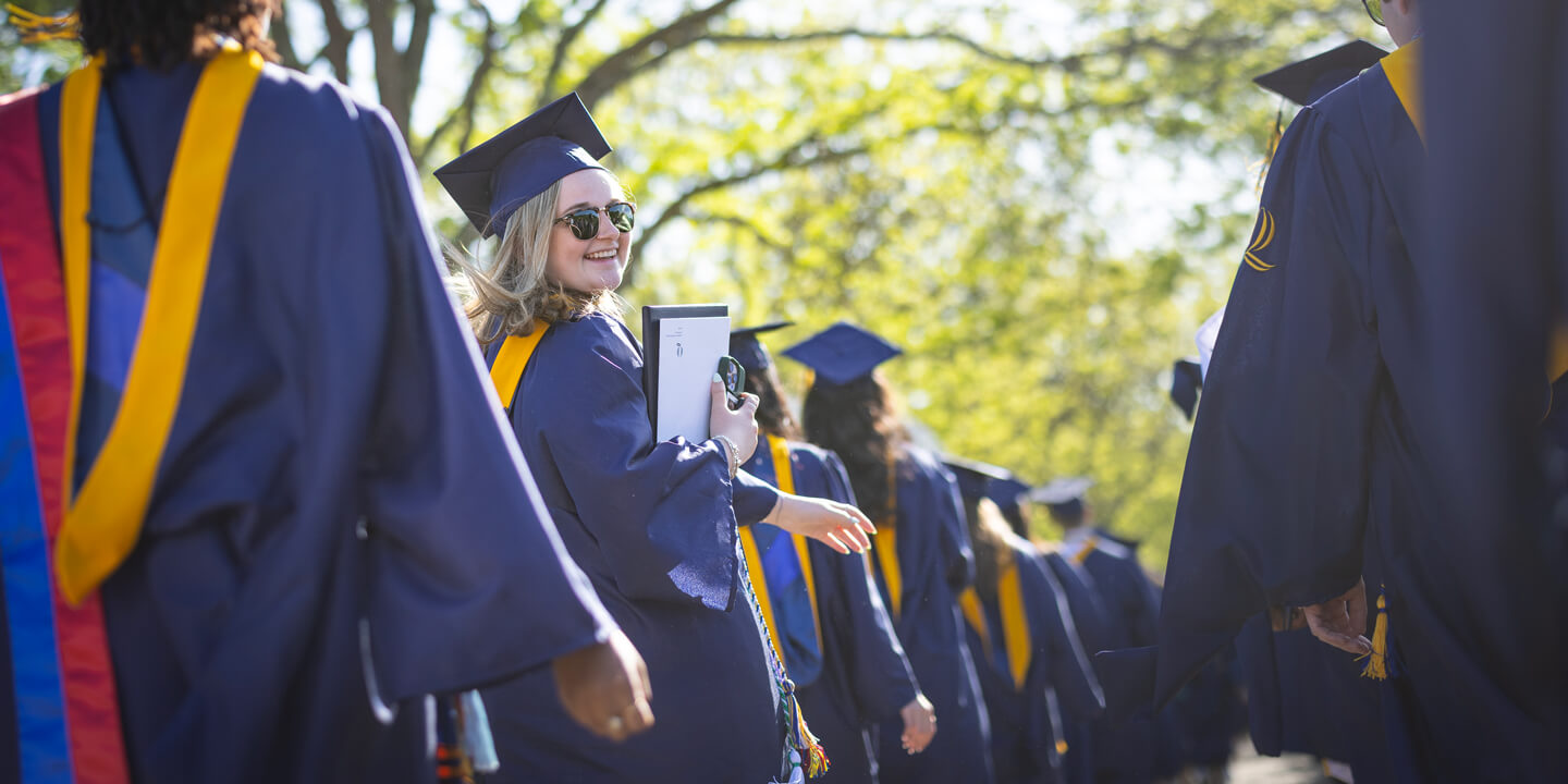 A graduate looks over her shoulder and smiles while walking in a line at Quinnipiac Commencement